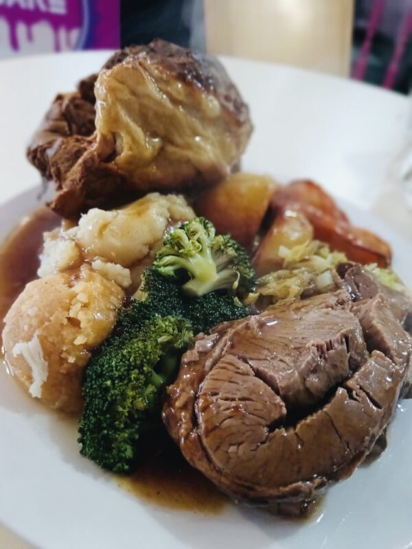 A plate of roast dinner featuring slices of tender beef covered in rich brown gravy, a golden Yorkshire pudding, mashed and roasted potatoes, steamed broccoli, and other vegetables. The meal is served on a white plate, with a blurred background including a hint of a pink and purple sign.