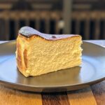 Current image: A slice of Basque burnt cheesecake with a golden-brown caramelized top and creamy, dense interior, served on a dark gray plate. The plate sits on a wooden table, with a blurred background featuring dark tones.