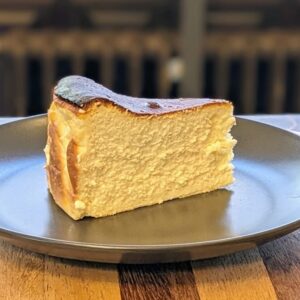 A slice of Basque burnt cheesecake with a golden-brown caramelized top and creamy, dense interior, served on a dark gray plate. The plate sits on a wooden table, with a blurred background featuring dark tones.