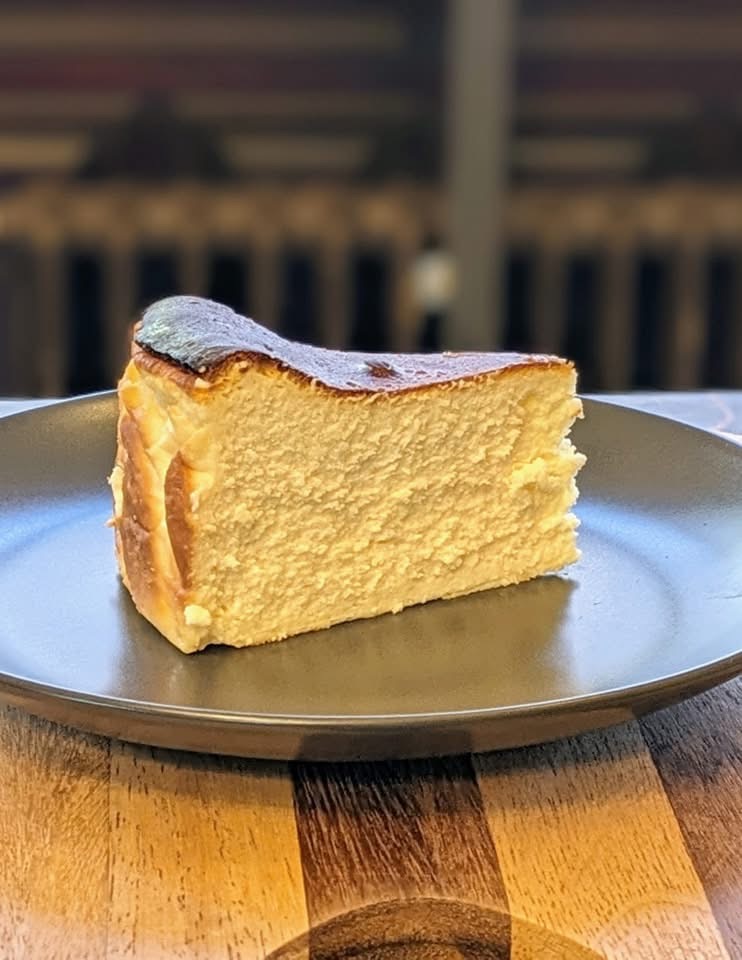 A slice of Basque burnt cheesecake with a golden-brown caramelized top and creamy, dense interior, served on a dark gray plate. The plate sits on a wooden table, with a blurred background featuring dark tones.