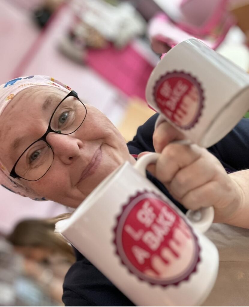 A person holds two mugs with the logo of 'L of a Bake' in a colorful café setting.